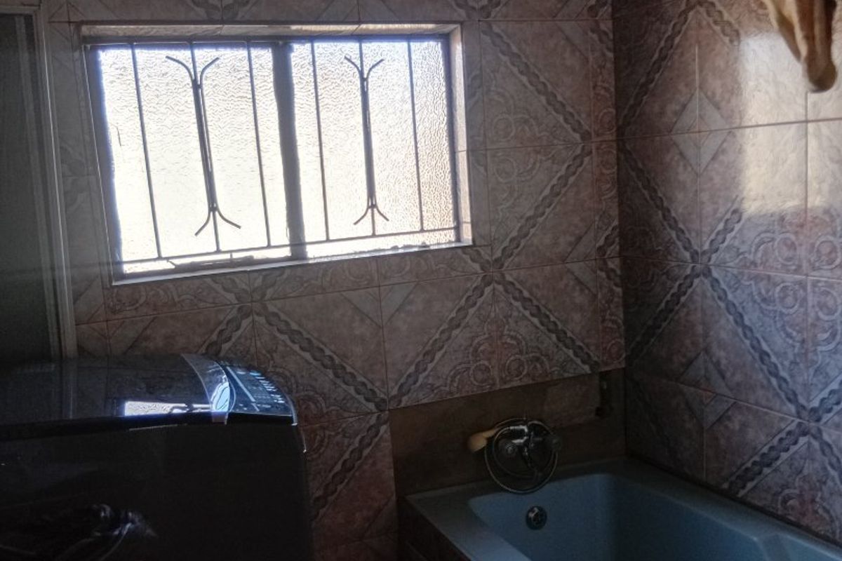 A dated bathroom featuring full-height patterned wall tiles, a built-in bathtub, and exposed timber ceiling beams with a window security grille.
