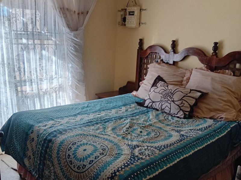 A bedroom featuring security bars on the window and a wall-mounted prepaid electricity meter, showing signs of ceiling water damage and worn flooring.