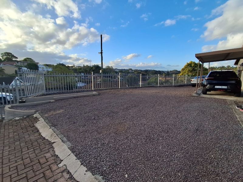A gravel driveway area featuring a carport, metal security fencing, and paved borders, offering an open outdoor space with neighborhood views.