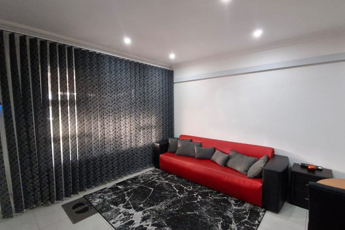 A well-lit living room featuring neutral tiled flooring, recessed ceiling downlights, and floor-to-ceiling vertical blinds.