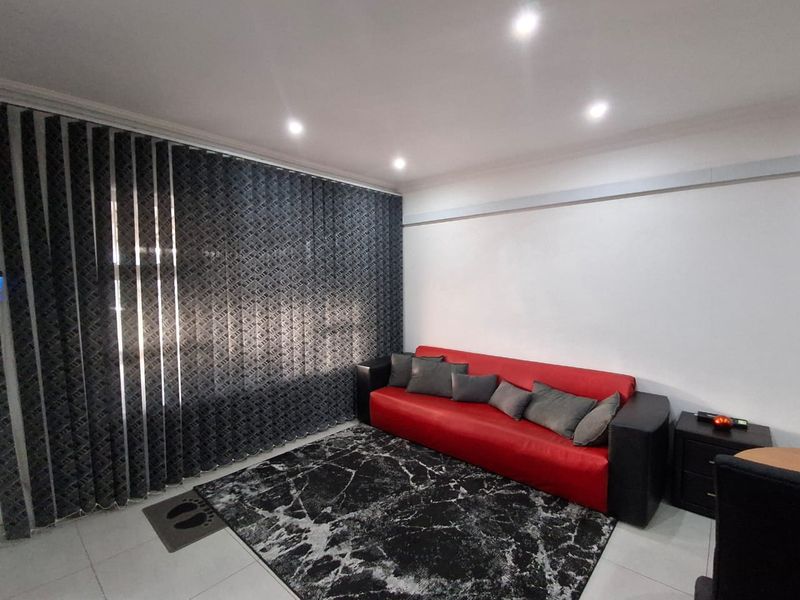 A well-lit living room featuring neutral tiled flooring, recessed ceiling downlights, and floor-to-ceiling vertical blinds.