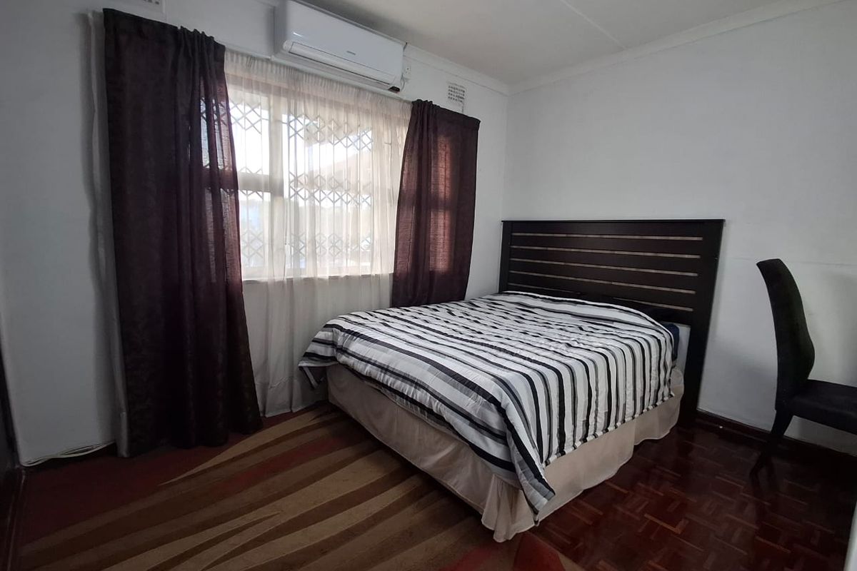 A bedroom featuring parquet flooring, a wall-mounted air conditioning unit, and a window fitted with a security gate.