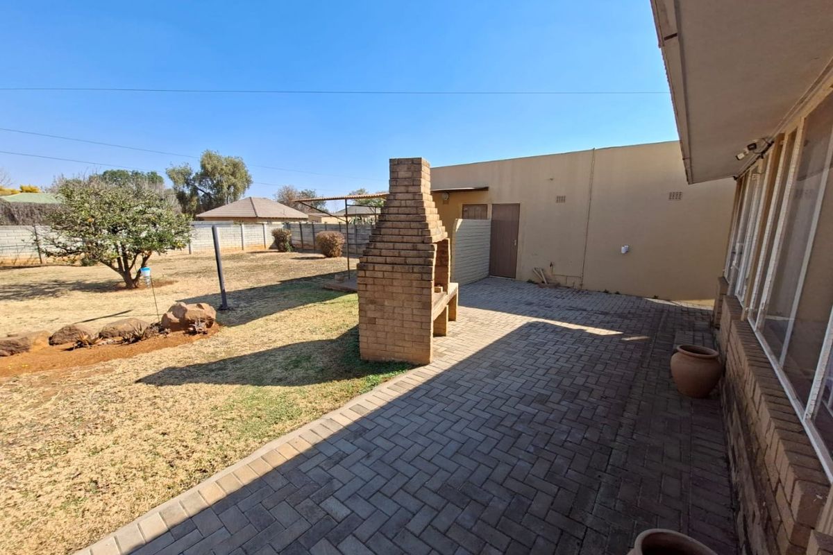 A spacious rear garden featuring a paved patio area with a built-in brick braai, overlooking a large lawn and perimeter wall.