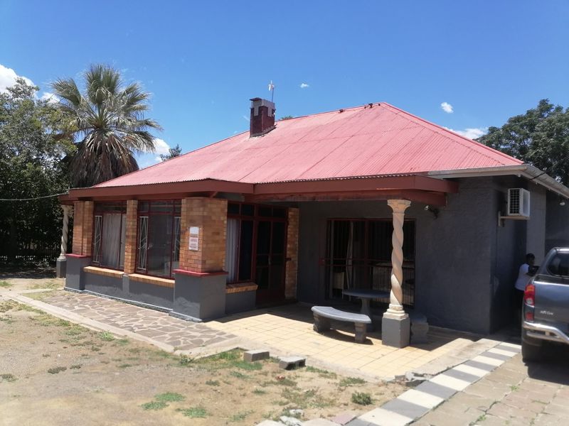 Front view of a single-story house featuring a red corrugated metal roof, a covered patio with a decorative column, and paved outdoor areas.