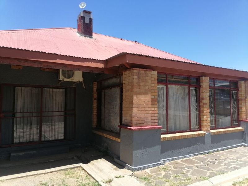 Front exterior view of a house featuring a corrugated metal roof, brick pillars, and stone paving, showing signs of exterior wear.