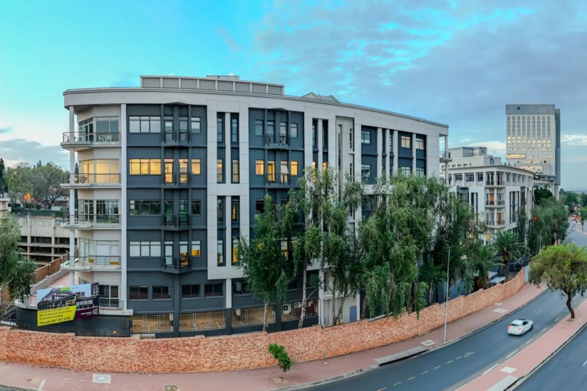 A panoramic view of a modern multi-story commercial building featuring a contemporary facade, balconies, and a secure perimeter brick wall along the street.