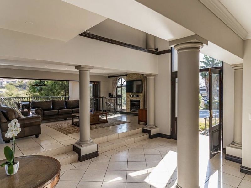 Spacious open-plan living area featuring a double-volume ceiling, decorative columns, tiled flooring, and a built-in fireplace with stone surround, leading to an outdoor balcony.