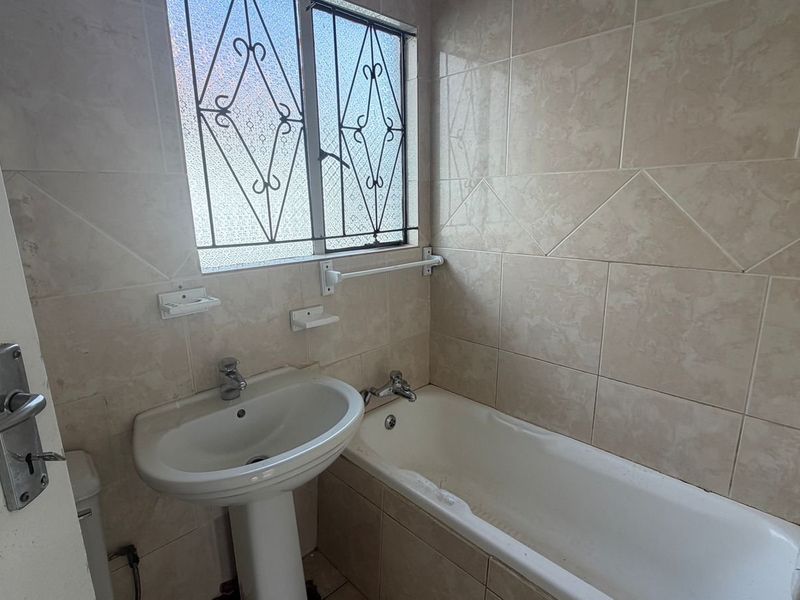 A functional bathroom featuring a pedestal basin, bathtub, and neutral wall tiling, complete with security bars on the window.