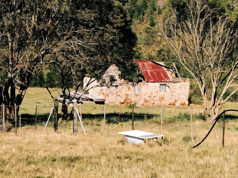 A dilapidated stone farm building with a partially collapsed corrugated metal roof, situated in a rural field with wire fencing.