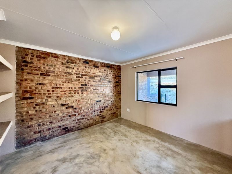 A bright, empty bedroom featuring a striking exposed brick feature wall, polished concrete floors, and a modern black-framed window.