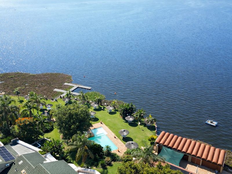 Aerial view of a waterfront property featuring a swimming pool, landscaped garden, private jetty, and solar panels on the main building roof.