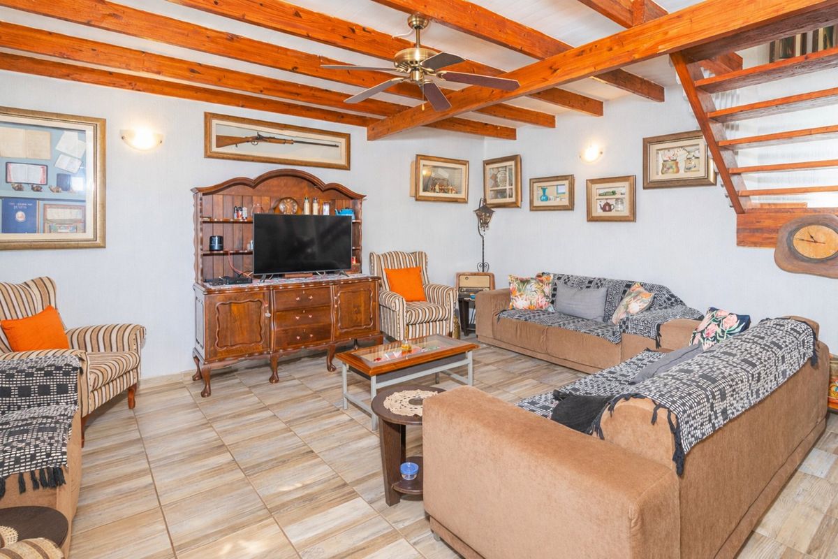 A living room featuring exposed timber ceiling beams, a ceiling fan, and tiled flooring, with an adjacent staircase leading to an upper level.