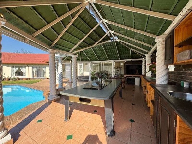 Spacious covered patio featuring a built-in braai, outdoor kitchen area with sink, and decorative columns, overlooking the swimming pool.