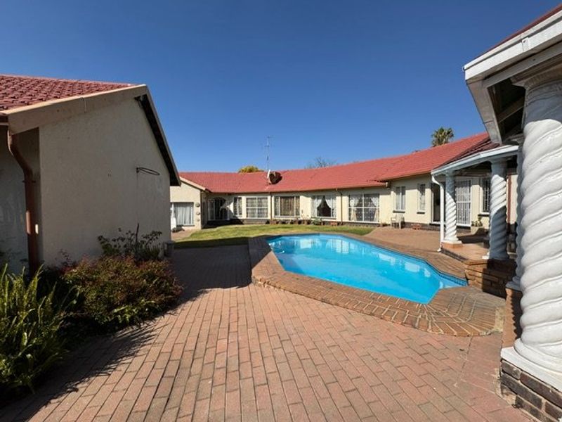A spacious outdoor pool area featuring a swimming pool, brick paved patio, and covered veranda, set against the rear facade of the house.