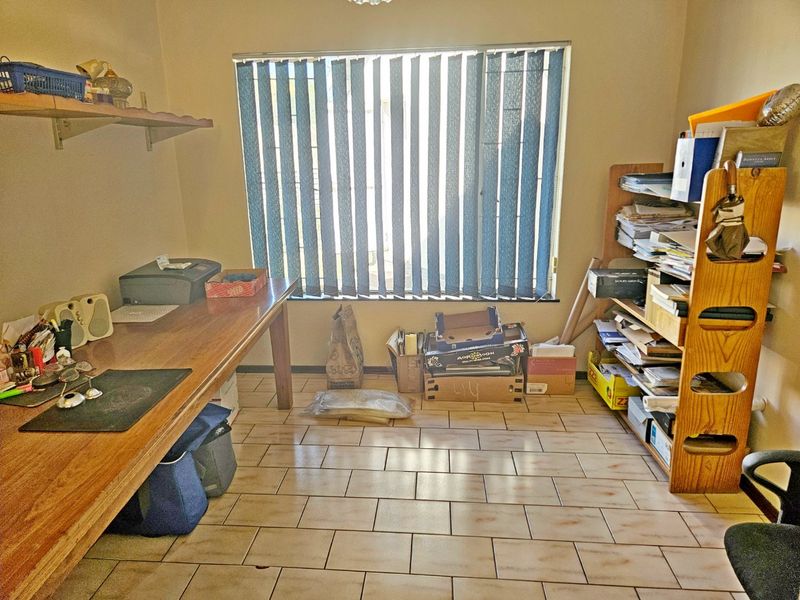 A functional home office space featuring ceramic tile flooring, a built-in desk, wall-mounted shelving, and vertical window blinds.