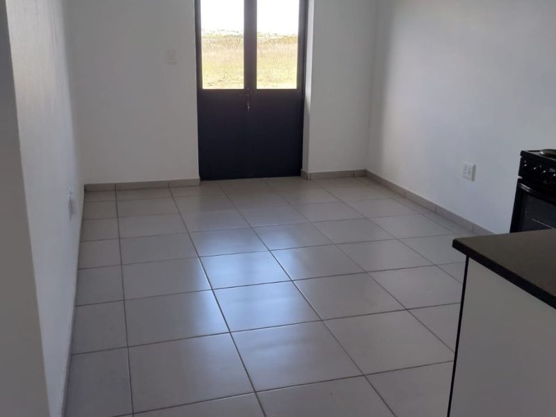 An empty open-plan living and kitchen area featuring neutral tiled flooring and a double door leading to the exterior.