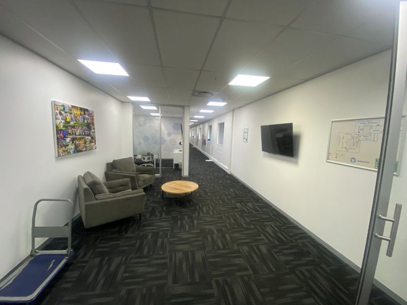 A long commercial hallway featuring carpeted flooring, suspended ceiling with recessed lighting, and glass partition walls leading to office spaces.