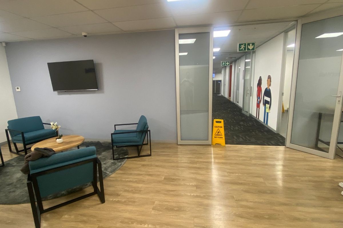 A commercial reception or waiting area featuring wood-look laminate flooring, glass partition doors leading to a carpeted hallway, and recessed ceiling lighting.