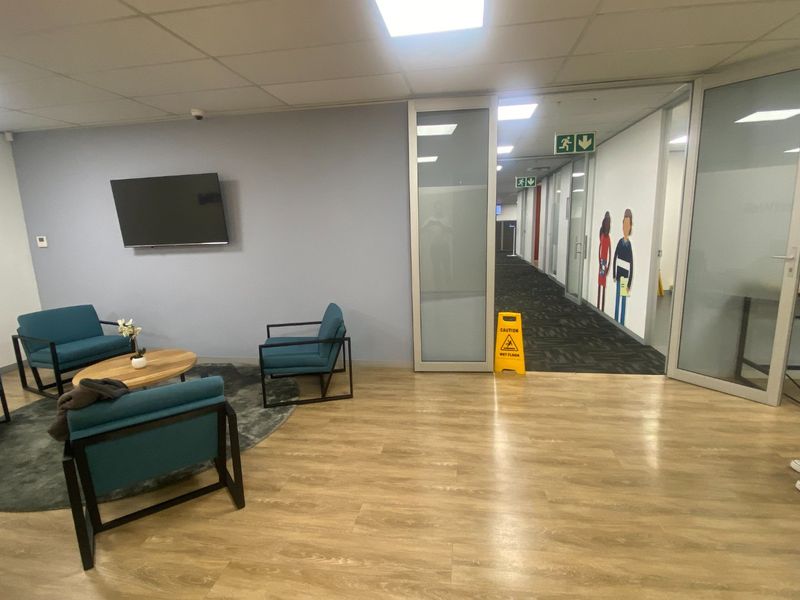 A commercial reception or waiting area featuring wood-look laminate flooring, glass partition doors leading to a carpeted hallway, and recessed ceiling lighting.