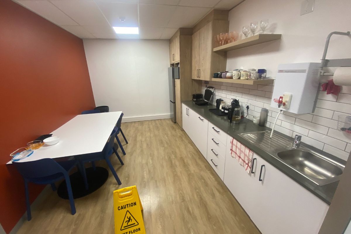 A functional commercial kitchenette featuring modern white cabinetry, wood-look flooring, subway tile backsplash, and an integrated instant hot water dispenser.