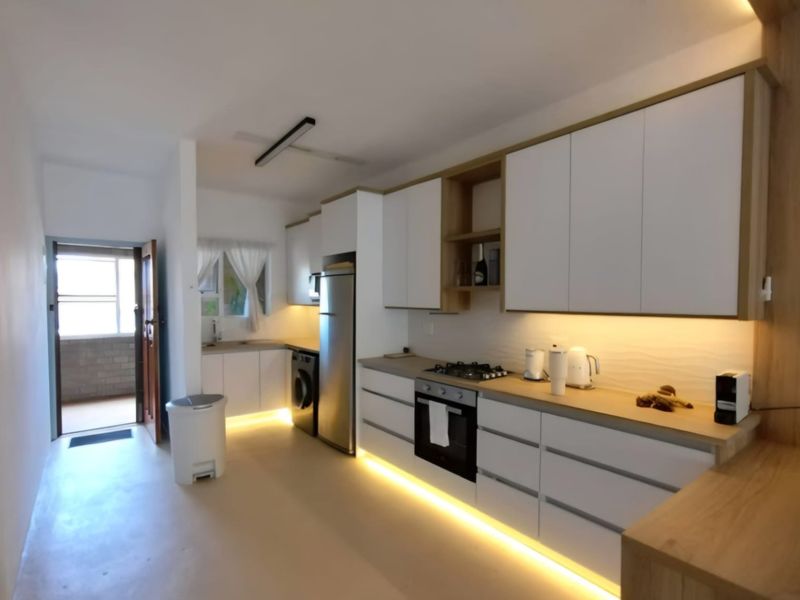 Modern kitchen featuring sleek white cabinetry, wood-tone accents, under-cabinet LED lighting, built-in oven, and gas hob, with direct access to an outdoor balcony area.