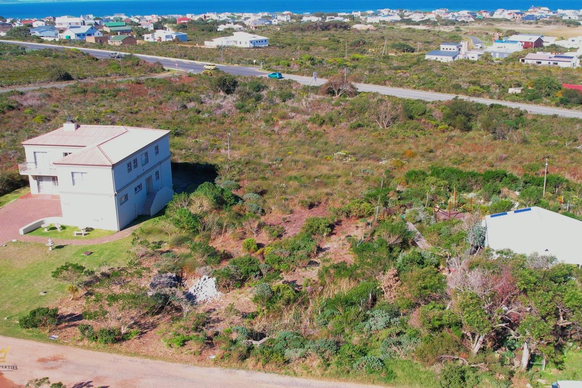 Aerial view of a vacant residential plot featuring natural vegetation, adjacent to an existing dwelling and paved road access.
