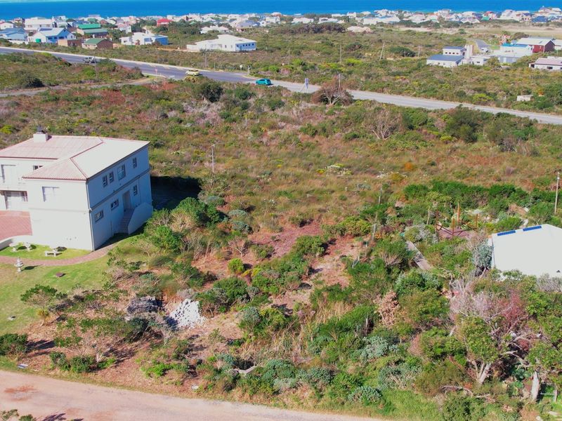 Aerial view of a vacant residential plot featuring natural vegetation, adjacent to an existing dwelling and paved road access.