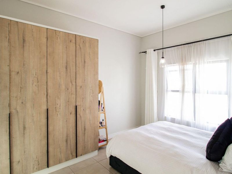 A well-maintained bedroom featuring modern wood-finish built-in cupboards, neutral tiled flooring, and natural light through a window with curtain fixtures.