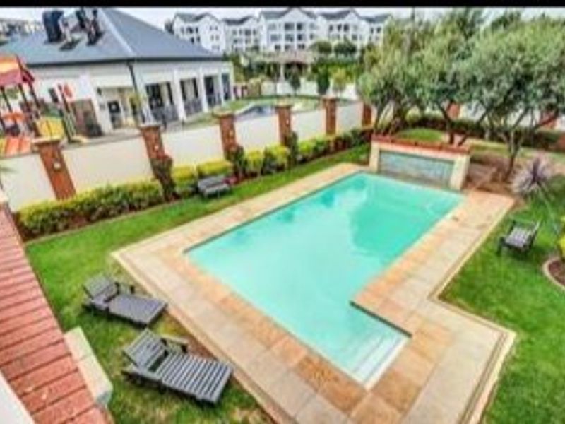 An elevated view of a communal swimming pool area featuring a landscaped garden, paved surround, and a decorative water feature.