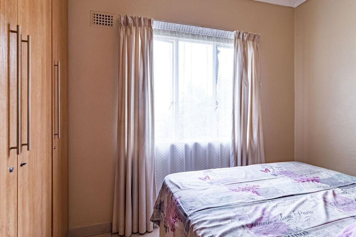 A bedroom featuring a built-in wooden wardrobe and a window with curtains, providing a functional and neutral living space.