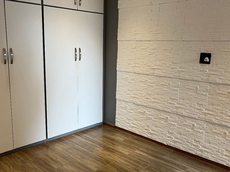 A clean, empty bedroom featuring wood-look tile flooring, ample built-in cupboard storage, and a modern white textured stone feature wall.