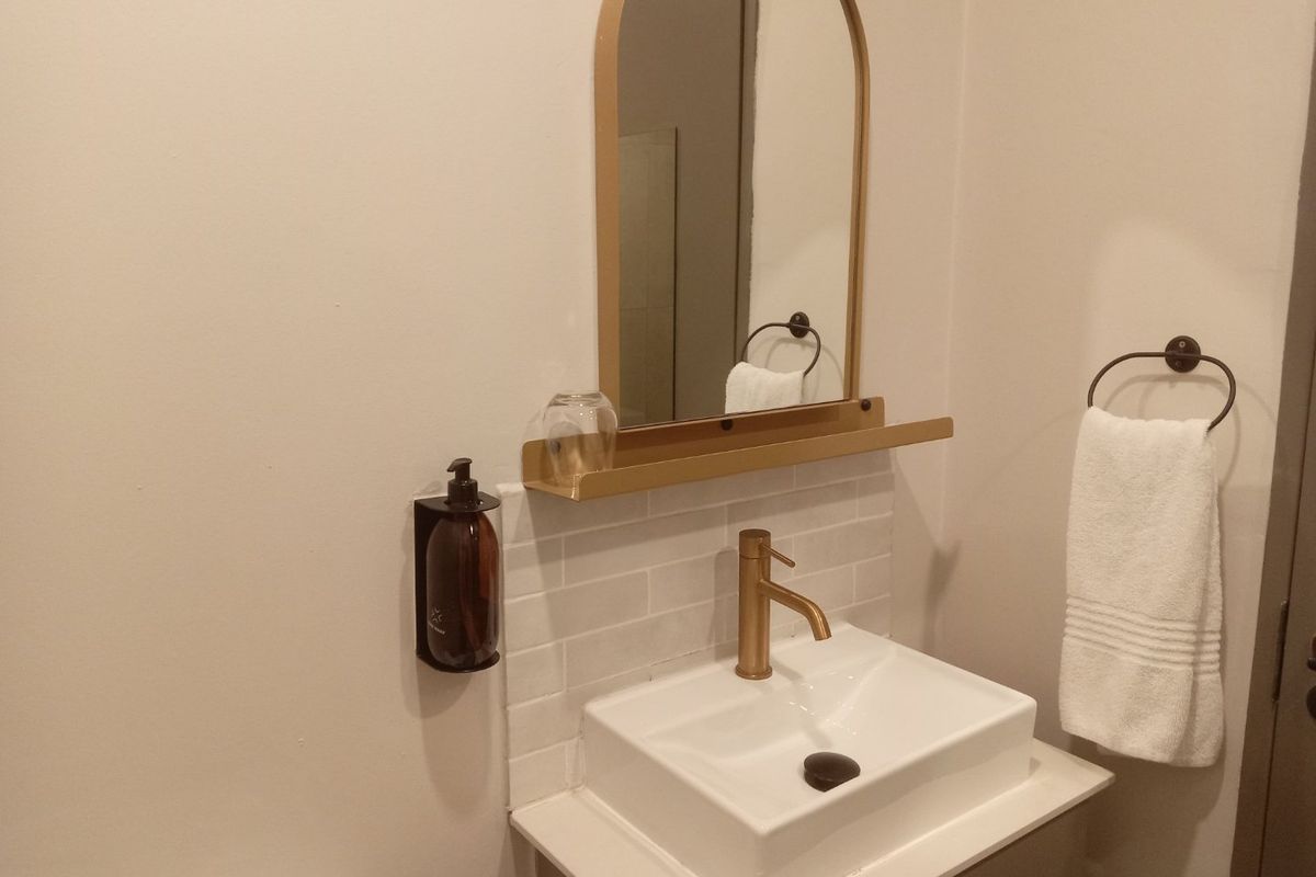 A modern bathroom vanity area featuring a white basin, brass faucet, subway tile backsplash, and an arched mirror with a built-in shelf.