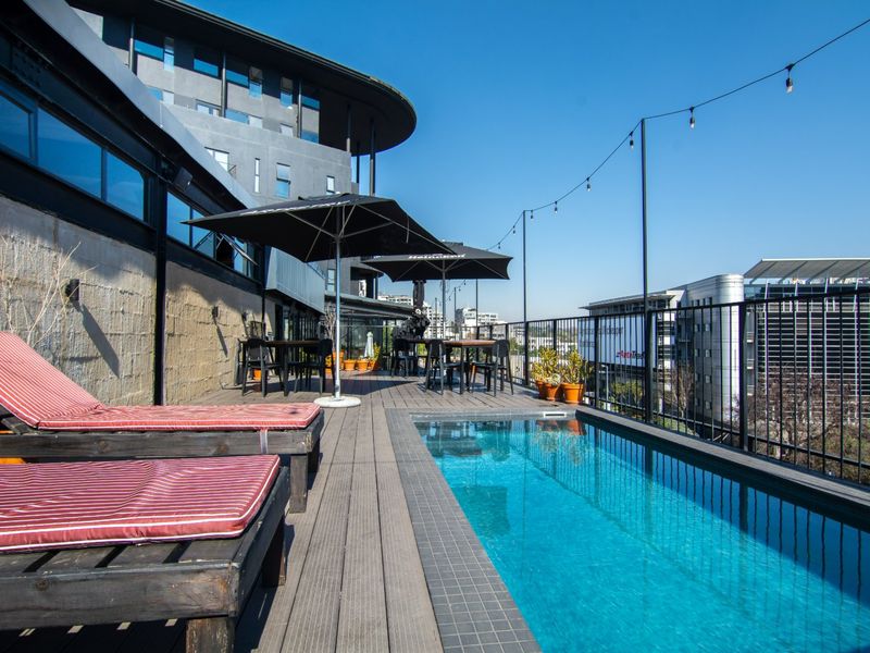 A rooftop terrace featuring a swimming pool, timber decking, and metal safety railings with city views.