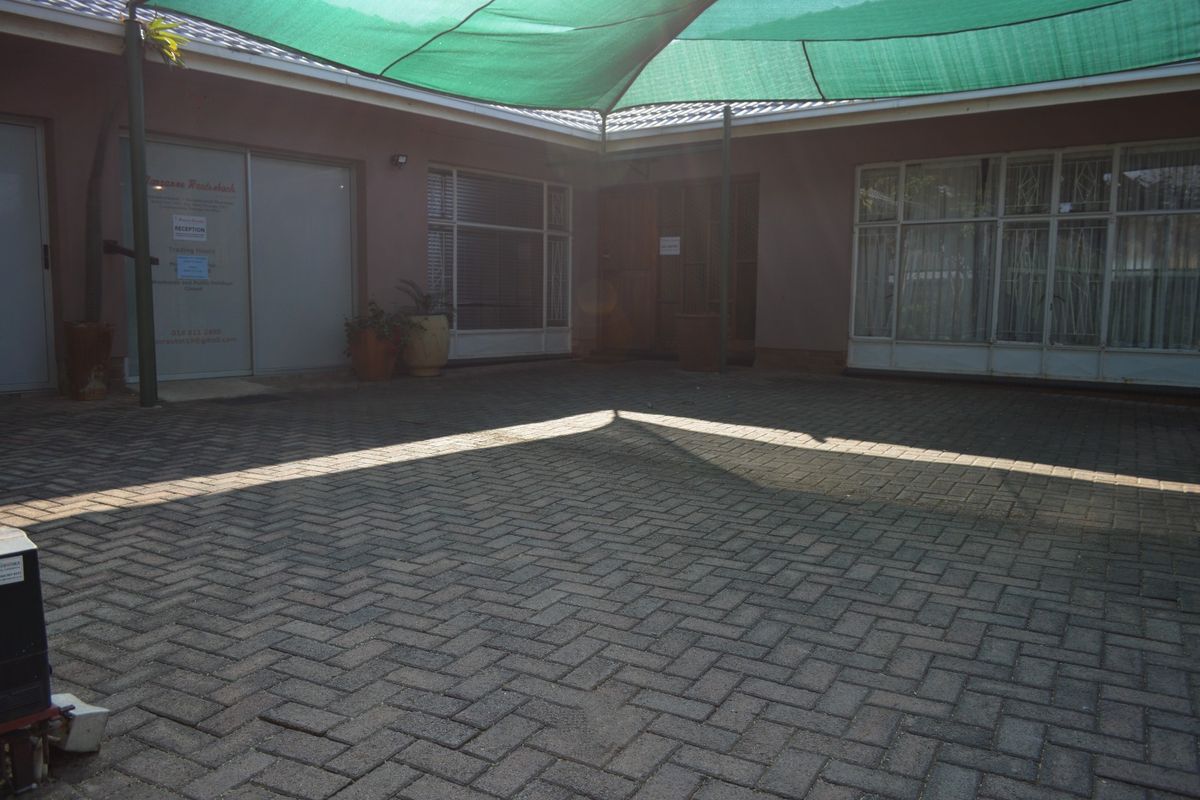 A paved rear courtyard area featuring a large overhead shade sail and access to the main building structure via multiple entry points.