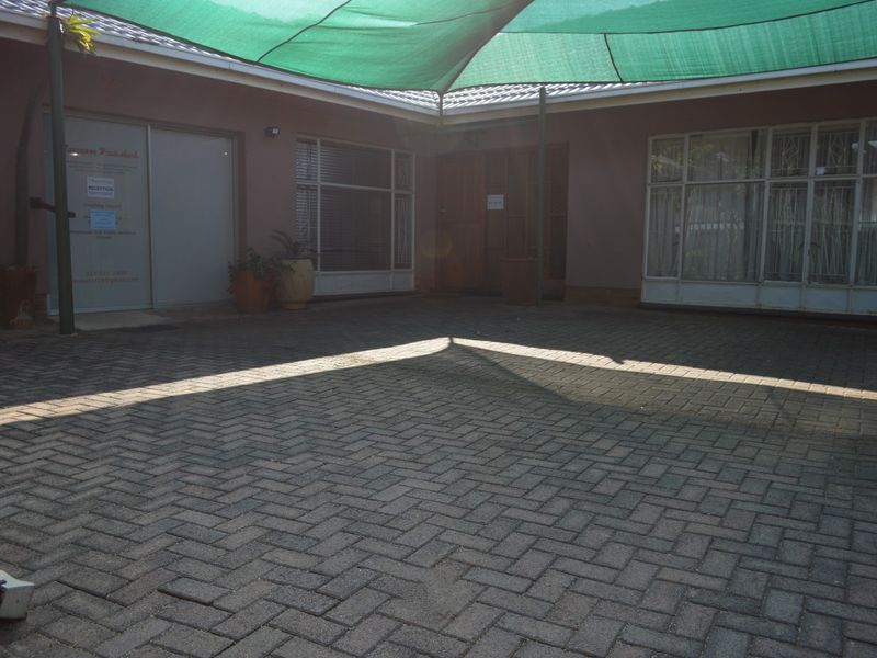 A paved rear courtyard area featuring a large overhead shade sail and access to the main building structure via multiple entry points.