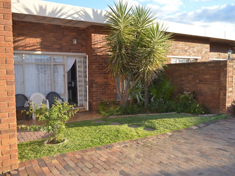 Front view of a townhouse featuring a brick facade, paved driveway, small garden area, and a secure entrance with a sliding glass door.