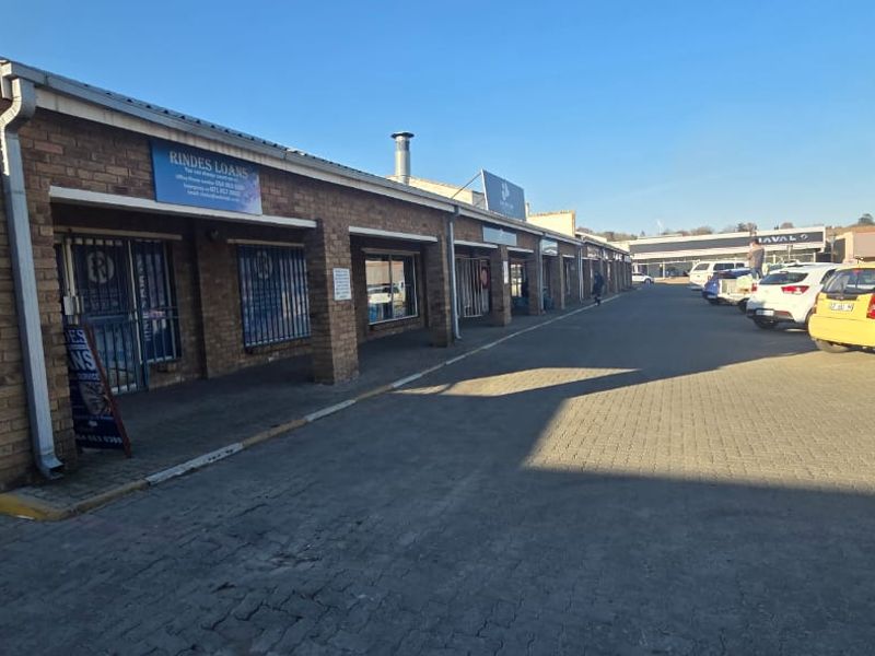 Exterior view of a commercial strip mall featuring face brick construction, shopfronts with security gates, and an adjacent paved parking area.