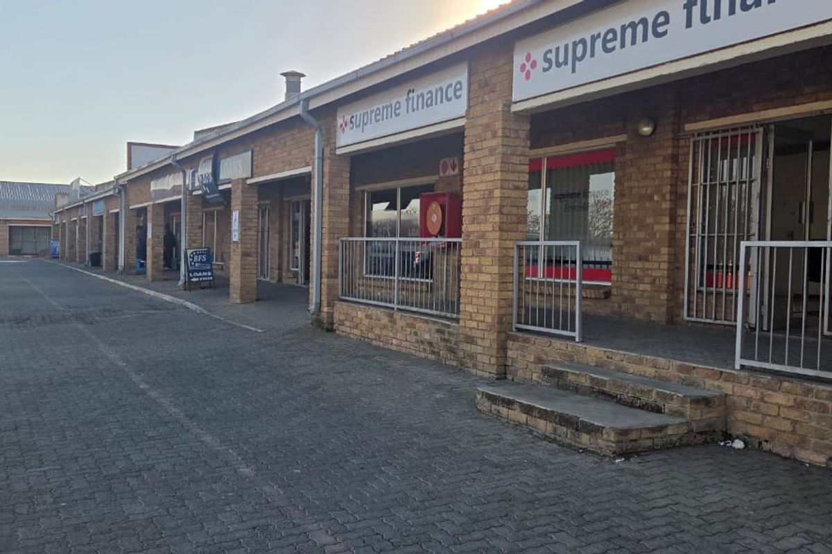 Exterior view of a commercial strip mall featuring face brick construction, paved parking area, and multiple storefront entrances with security gates.