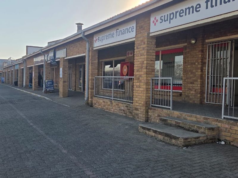 Exterior view of a commercial strip mall featuring face brick construction, paved parking area, and multiple storefront entrances with security gates.