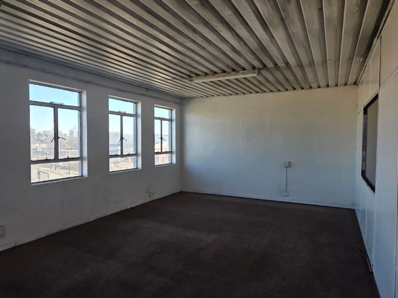 An empty, functional office space featuring large windows for natural light, a corrugated metal ceiling, and carpeted flooring.
