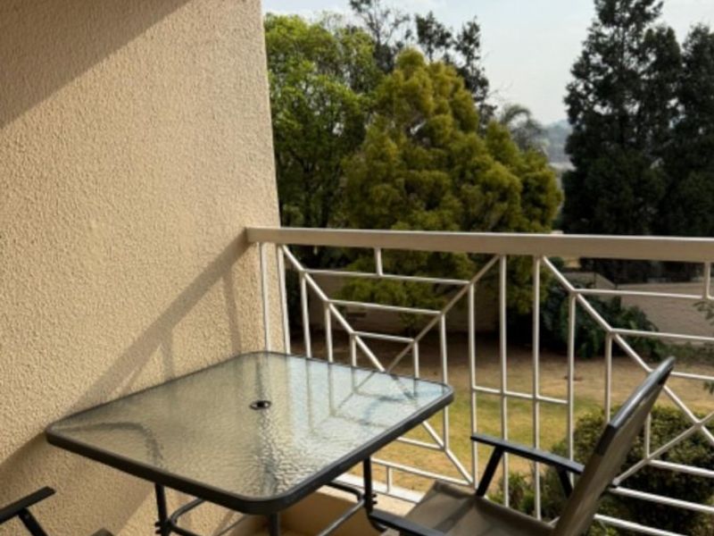 A covered balcony featuring tiled flooring and a white metal balustrade, offering an elevated view of the surrounding trees and garden area.