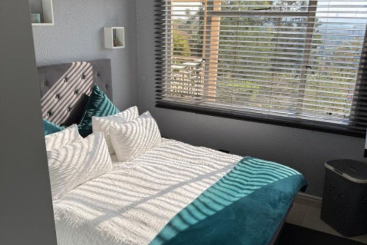 A cozy bedroom featuring grey walls, crown molding, a ceiling fan, and large windows with venetian blinds offering natural light and outdoor views.