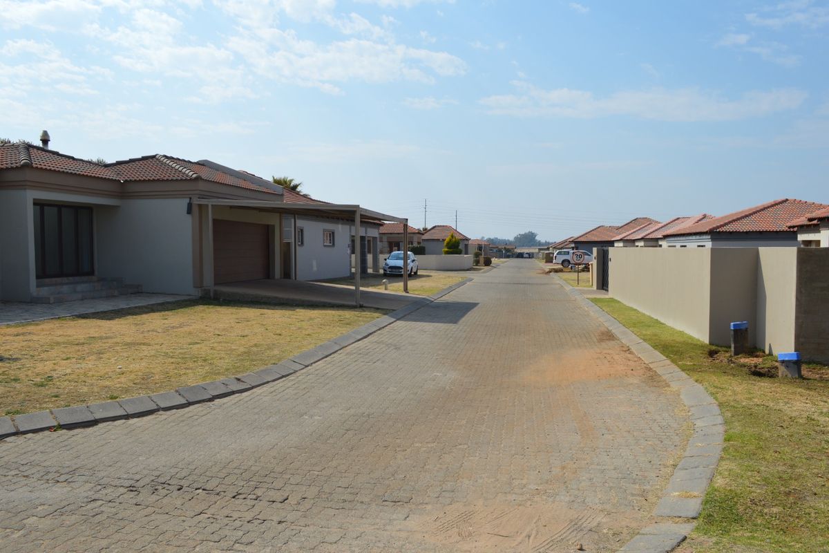 A view of a residential street in a housing estate featuring paved roads, boundary walls, and modern single-story homes with tiled roofs.