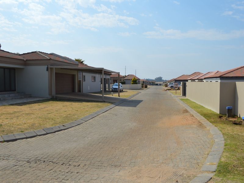 A view of a residential street in a housing estate featuring paved roads, boundary walls, and modern single-story homes with tiled roofs.