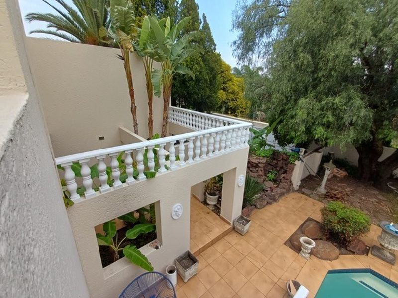 An elevated balcony with a classic balustrade overlooking a tiled patio area and swimming pool, surrounded by established garden greenery.