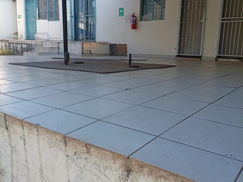 A covered outdoor patio area featuring tiled flooring, security gates, and a corrugated metal roof structure, showing signs of exterior wear.