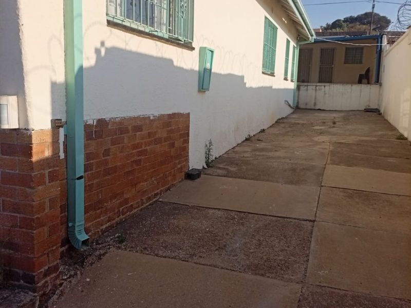 A side exterior view of the property featuring a paved walkway, brick and plaster facade, security bars on windows, and perimeter boundary walling.
