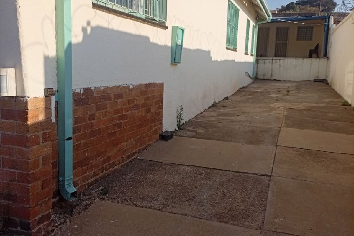 A side exterior view of the property featuring a paved walkway, security bars on windows, and a brick plinth along the building facade.
