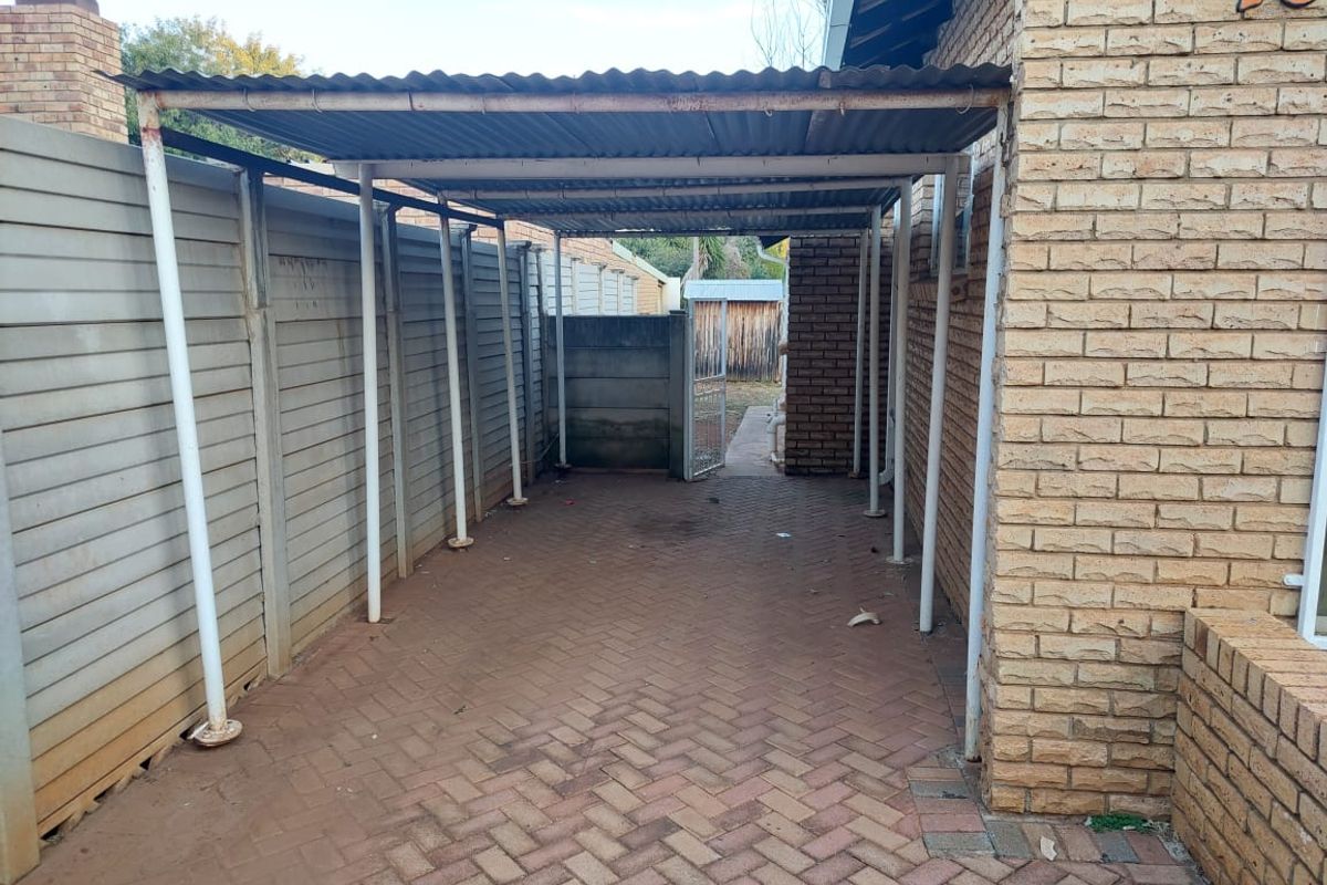 A covered outdoor walkway or carport area featuring brick paving, a corrugated metal roof, and precast concrete boundary walls.