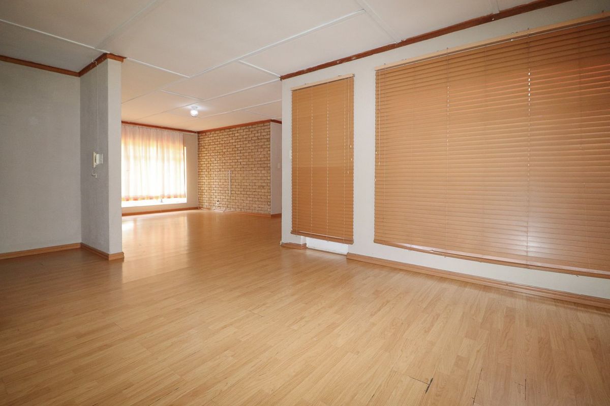 Spacious, empty living area featuring laminate flooring, an exposed brick feature wall, and large windows with fitted venetian blinds.